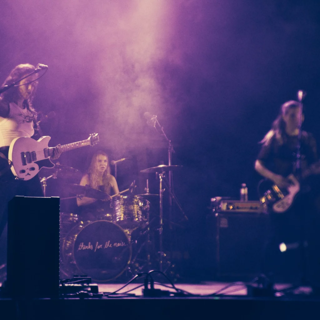 Photo d’un concert du groupe MADAM. Une chanteuse guitarise à gauche. Une bassiste à droite. Un batteuse au centre dans le fond. Dans de la fumée de couleur violette.