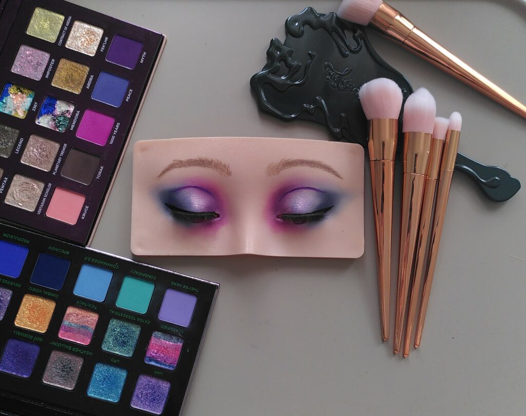 Visage d’entraînement au maquillage entouré de pinceaux, d'un miroir et de deux palettes ouvertes. Le maquillage est bleu, violet, lavande et rose.