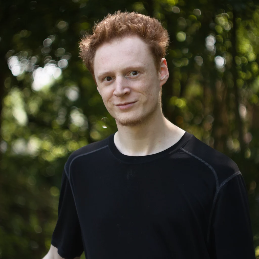 Photo d’un homme blanc et roux, avec un tshirt noir. Le fond flou est végétal et parsemé de bokeh
